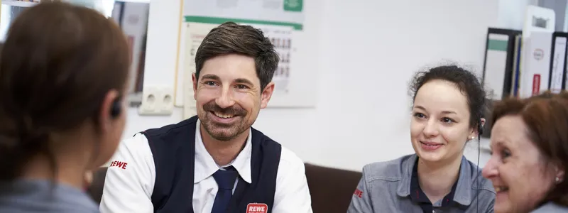 REWE Mitarbeitende sitzen gemeinsam an einem Tisch und unterhalten sich froehlich