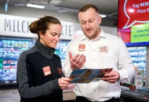 Zwei Rewe Mitarbeitende schauen gemeinsam auf ein Prospekt. 
