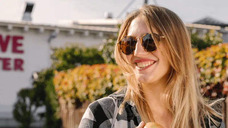 Eine Frau mit Sonnenbrille laechelt, Natur und ein Rewe Markt befinden sich im Hintergrund