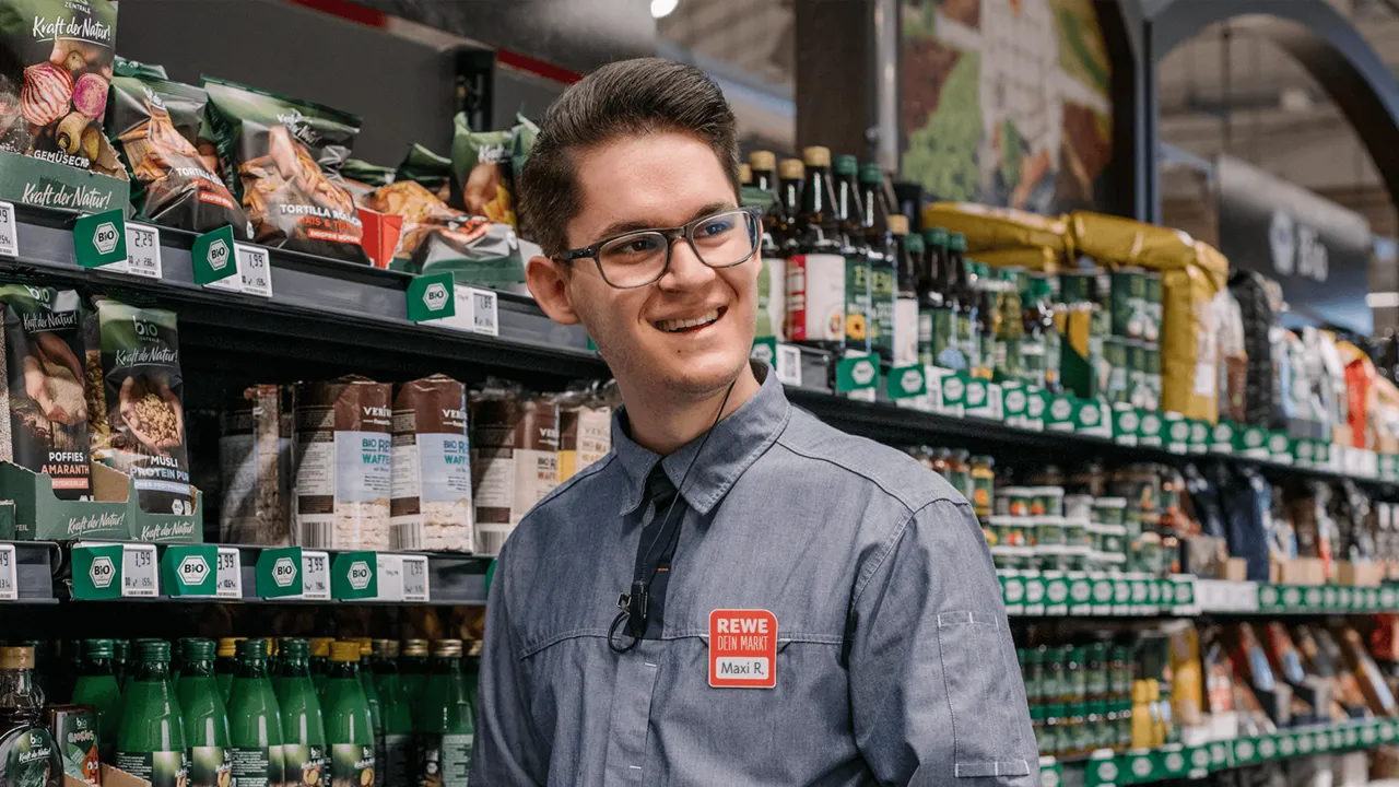 Ein REWE Student im Traineeprogramm zur Selbstaendigkeit als Kaufmann steht vor dem Olivenoel Regal