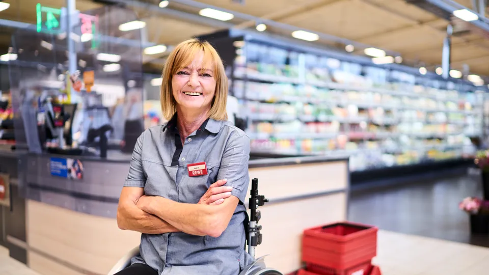 Eine Frau im Rollstuhl, traegt Rewe Arbeitskleidung und befindet sich in einer Rewe Filiale.