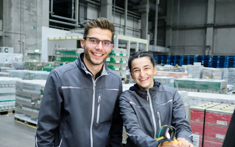 Zwei Mitarbeitende stehen gemeinsam in Arbeitskleidung im Logistik Lager