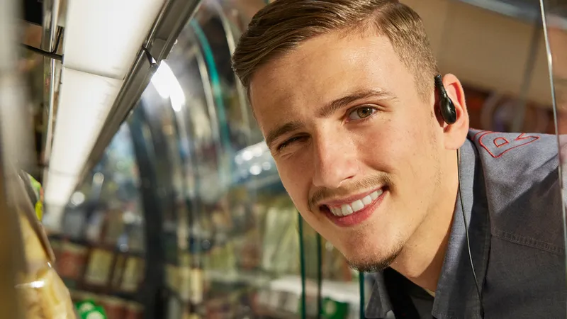 Ein Rewe Mitarbeiter mit einem Headset im Ohr laechelt.