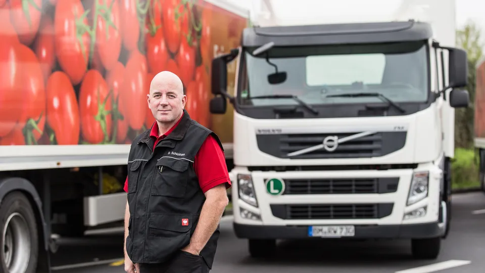 Ein REWE Logistik Mitarbeiter steht vor einem LKW und blickt direkt in die Kamera