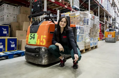 Eine REWE Auszubildende zur Fachkraft für Lagerlogistik posiert vor einem Transportwagen