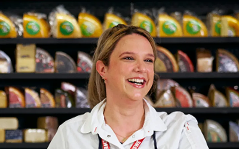 Eine lachende Rewe Mitarbeiterin steht hinter der Frischetheke vor einem großen Regal mit verschiedenen Käsesorten.