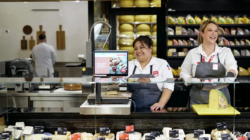 Zwei Mitarbeiterinnnen in REWE Arbeitskleidung bedienen freundlich an der Frischetheke mit verschiedenen Käsesorten vor einem großen Käseregal.